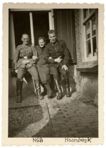 Zwart wit foto van twee duitste militaire met in het midden een oudere vrouw leunend op een vensterbank.